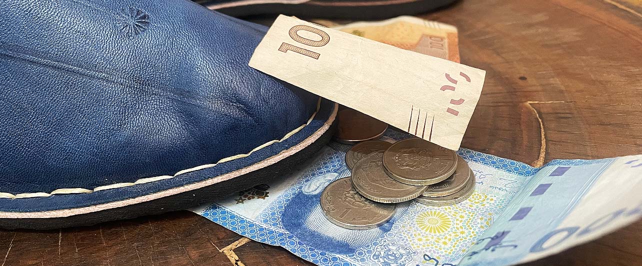 Blue leather babouches styled with Moroccan banknotes and an Artisanat Shop tag