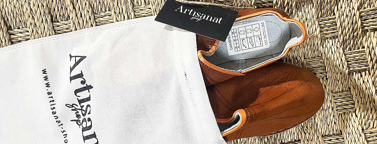 Brown leather babouches emerging from a white Artisanat Shop bag on woven straw